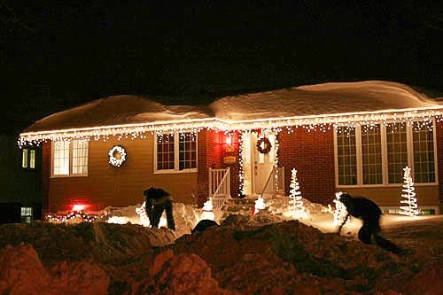 shovelling snow in front of a house decorated with christmas lights